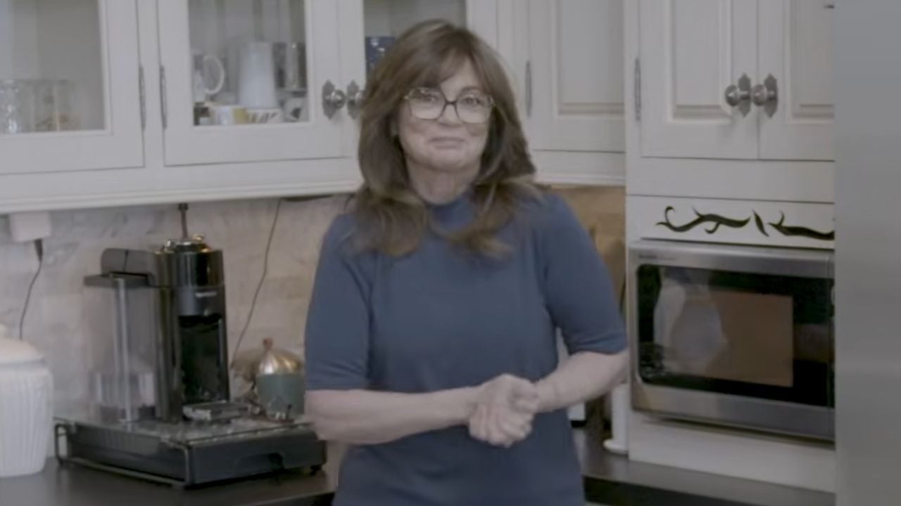 A woman with long brown hair and glasses stands in a kitchen, smiling slightly. Behind her are white cabinets, a microwave, a coffee machine, and a teapot.