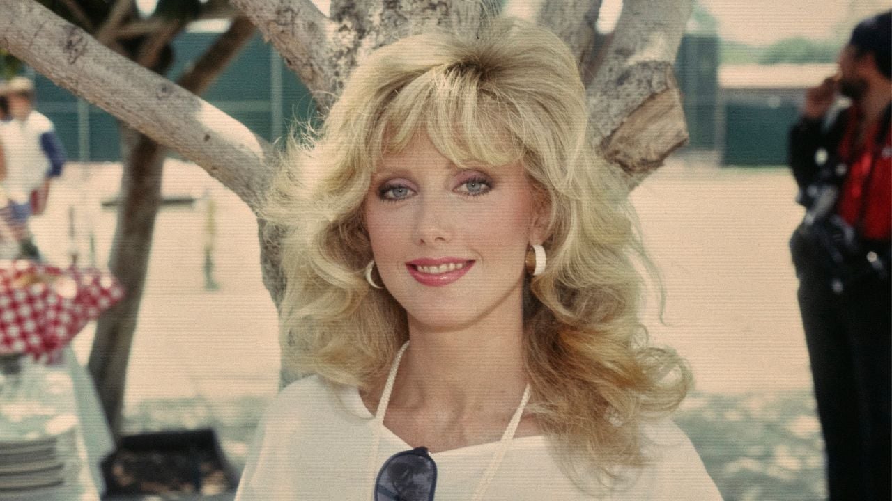 A medium shot of a smiling woman with voluminous, wavy blonde hair and blue eyes, wearing a white top and hoop earrings, standing in front of a tree outdoors. In the background, a person in a red shirt is partially visible.