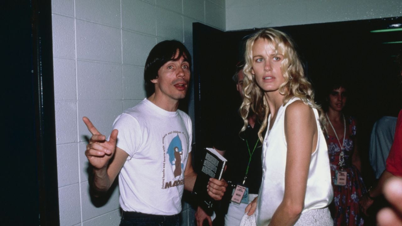 A man in a white t-shirt holding a book gestures while standing next to a woman with long blonde hair in a white outfit. They are in a hallway with people and lanyards in the background.