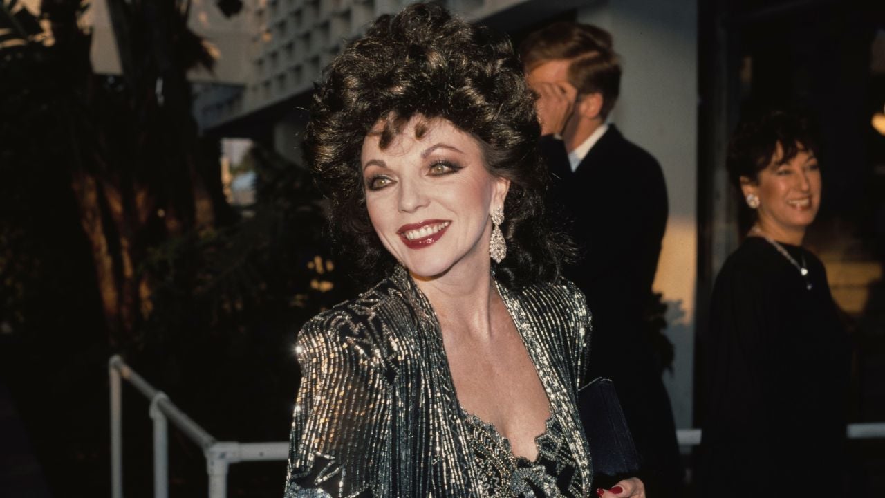 A woman in a shiny silver dress smiles at the camera outdoors. She has curly dark hair and wears bold makeup, earrings, and holds a black clutch. Other people are in the background.