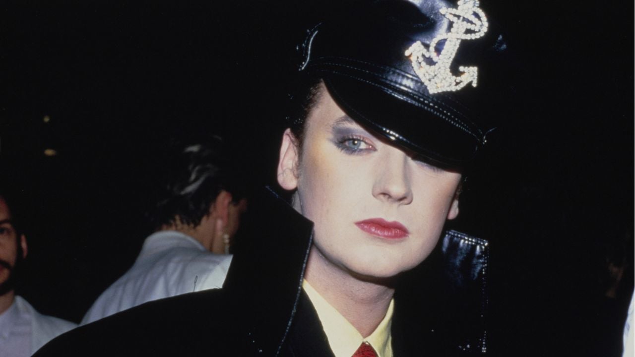 Person wearing dramatic makeup, a black leather hat with a rhinestone anchor, and a high-collared black coat. The background is dimly lit, with other people partially visible.