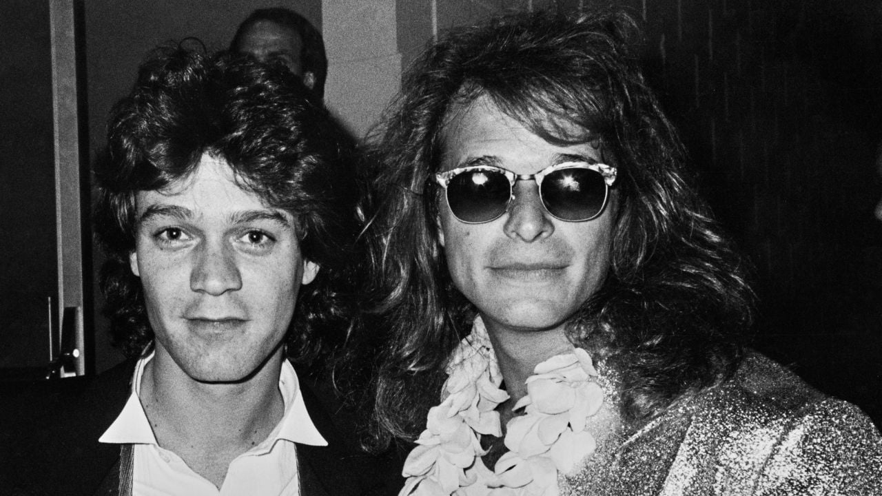 Two men in black and white, one with short dark hair wearing a collared shirt, the other with long hair, sunglasses, and a flower lei, stand close together and look at the camera.
