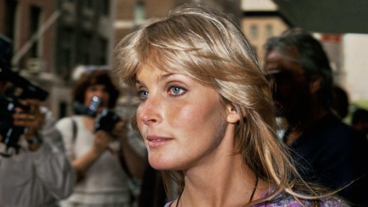 A woman with blonde hair and blue eyes walks outside in a city, looking ahead. People and photographers are in the background, holding cameras.