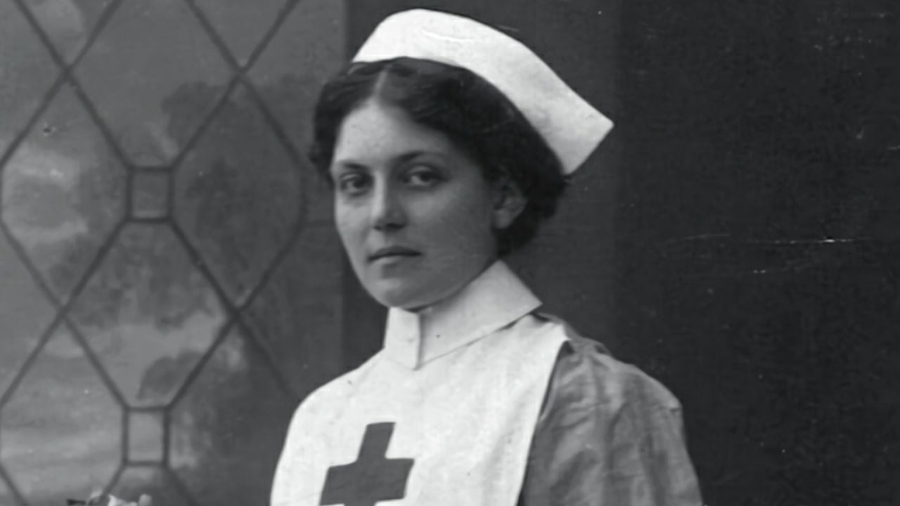 Black and white photo of a woman in a nurse’s uniform with a Red Cross symbol, standing in front of a window with a geometric pattern, looking directly at the camera.