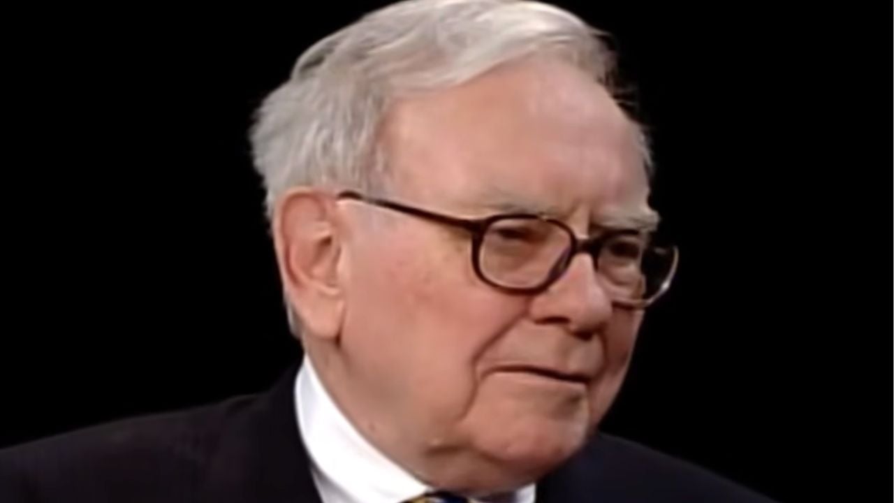 An older man with white hair and glasses, wearing a dark suit, light shirt, and tie, is looking slightly to the side against a dark background.