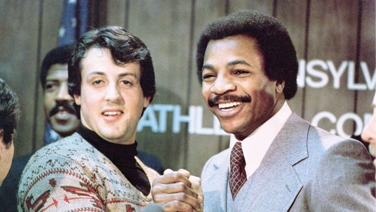 Two men smiling and clasping hands in front of a wooden wall with partially visible text. One man wears a patterned sweater, the other a light-colored suit and tie. Other people are blurred in the background.