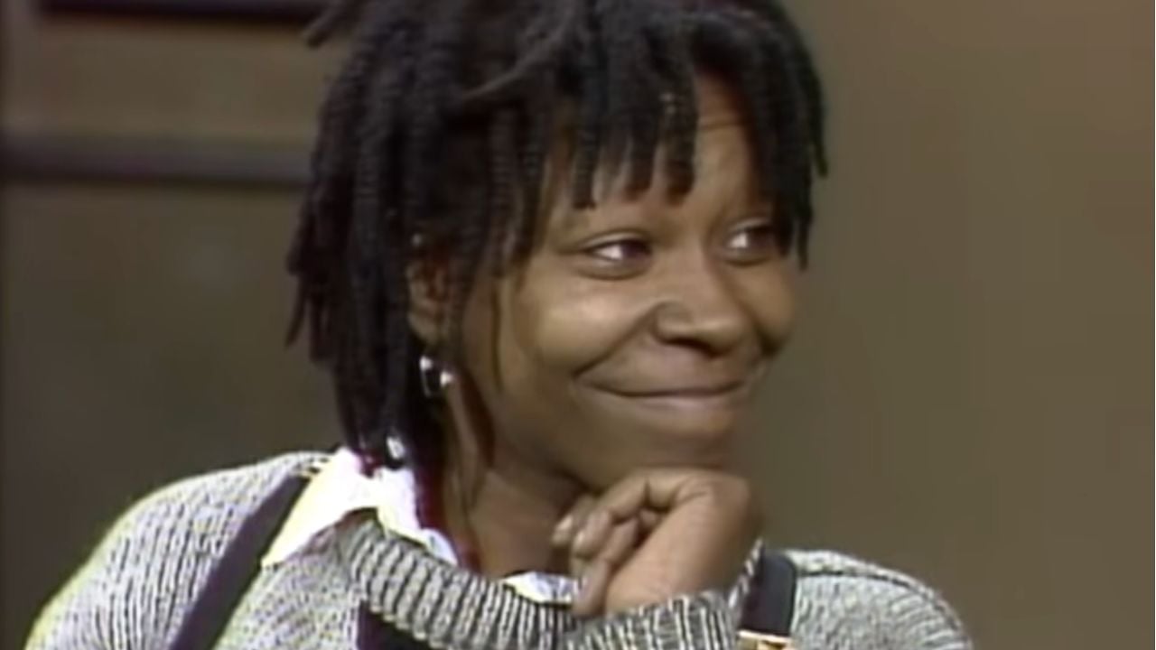 A person with short dreadlocks and a gray sweater smiles softly, resting their chin on their hand. The background is blurred.