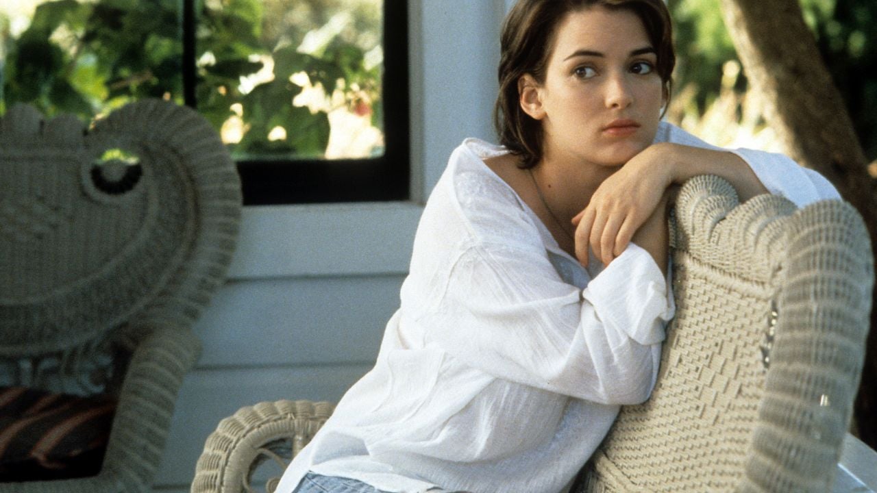 A young woman in a white shirt leans on a wicker chair, gazing thoughtfully into the distance. She sits outdoors near a window with greenery visible behind her.