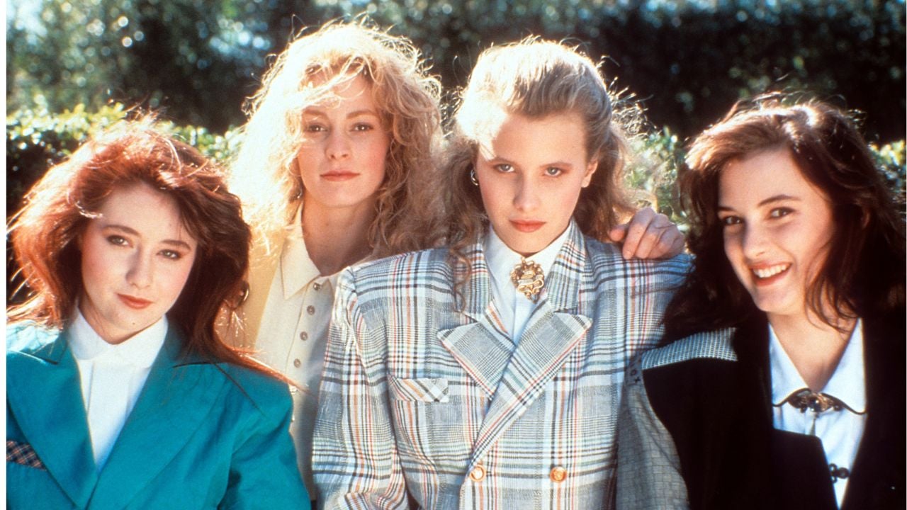 Four young women in 1980s fashion, with styled hair and bold blazers, stand together outside, smiling or posing seriously for the camera.
