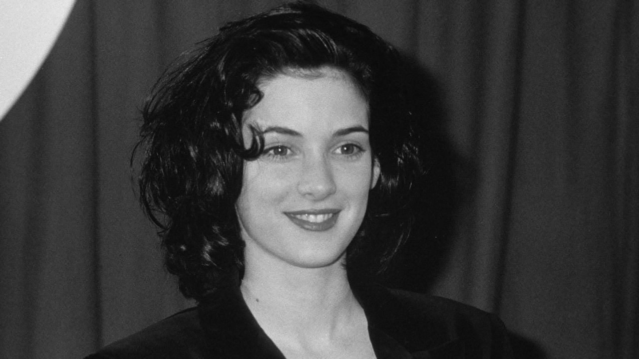A young woman with wavy dark hair, wearing a dark jacket, smiles in front of a curtain backdrop in this black-and-white photo.