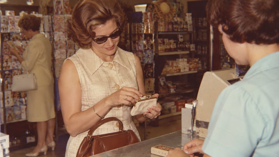 A woman in sunglasses pays at a store counter with a coin purse, while another woman in the background browses shelves filled with colorful merchandise.