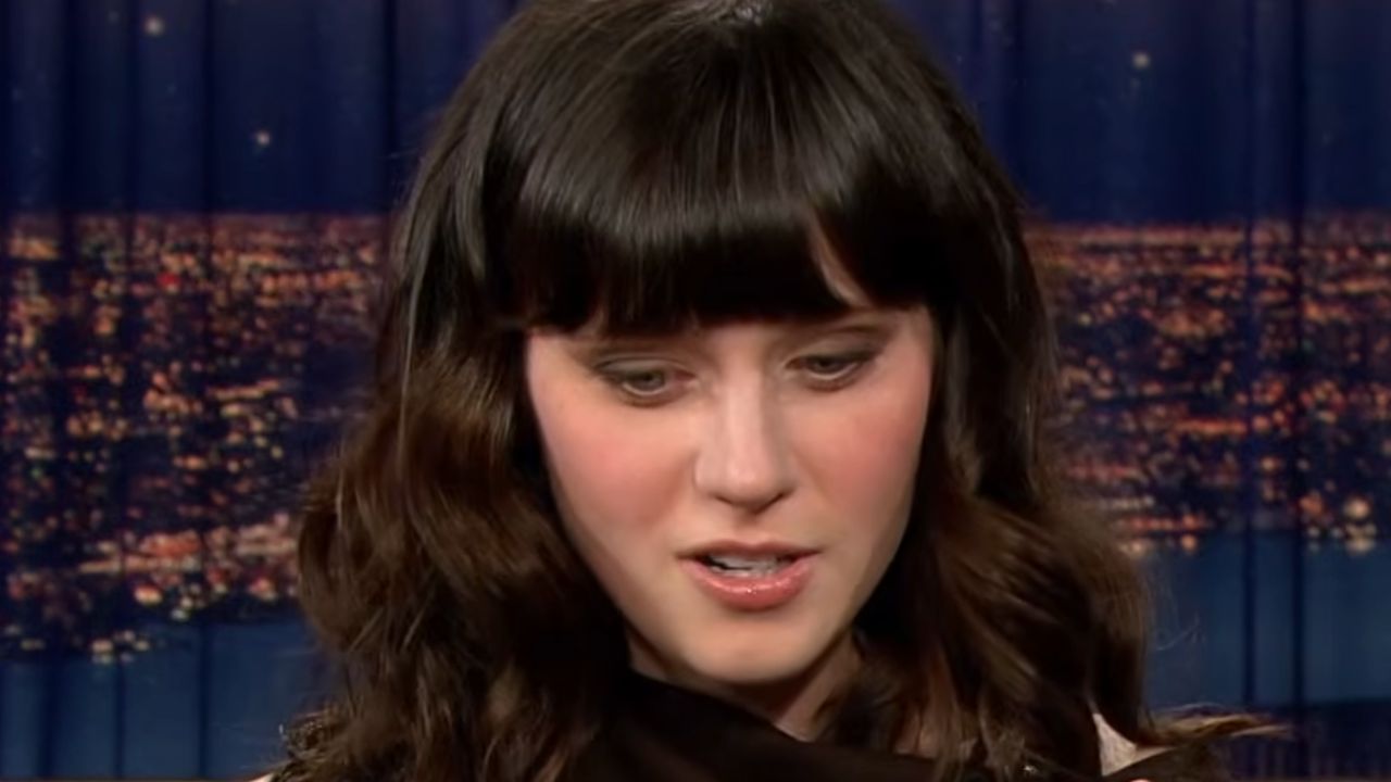 A woman with long dark wavy hair and bangs looks down, wearing a dark outfit. The blurry city lights are visible in the background.