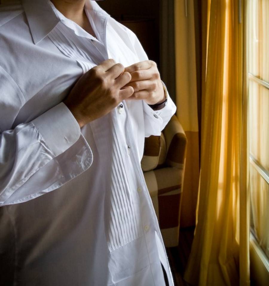 A person standing by a sunlit window buttons up a white dress shirt, with only their torso and hands visible.