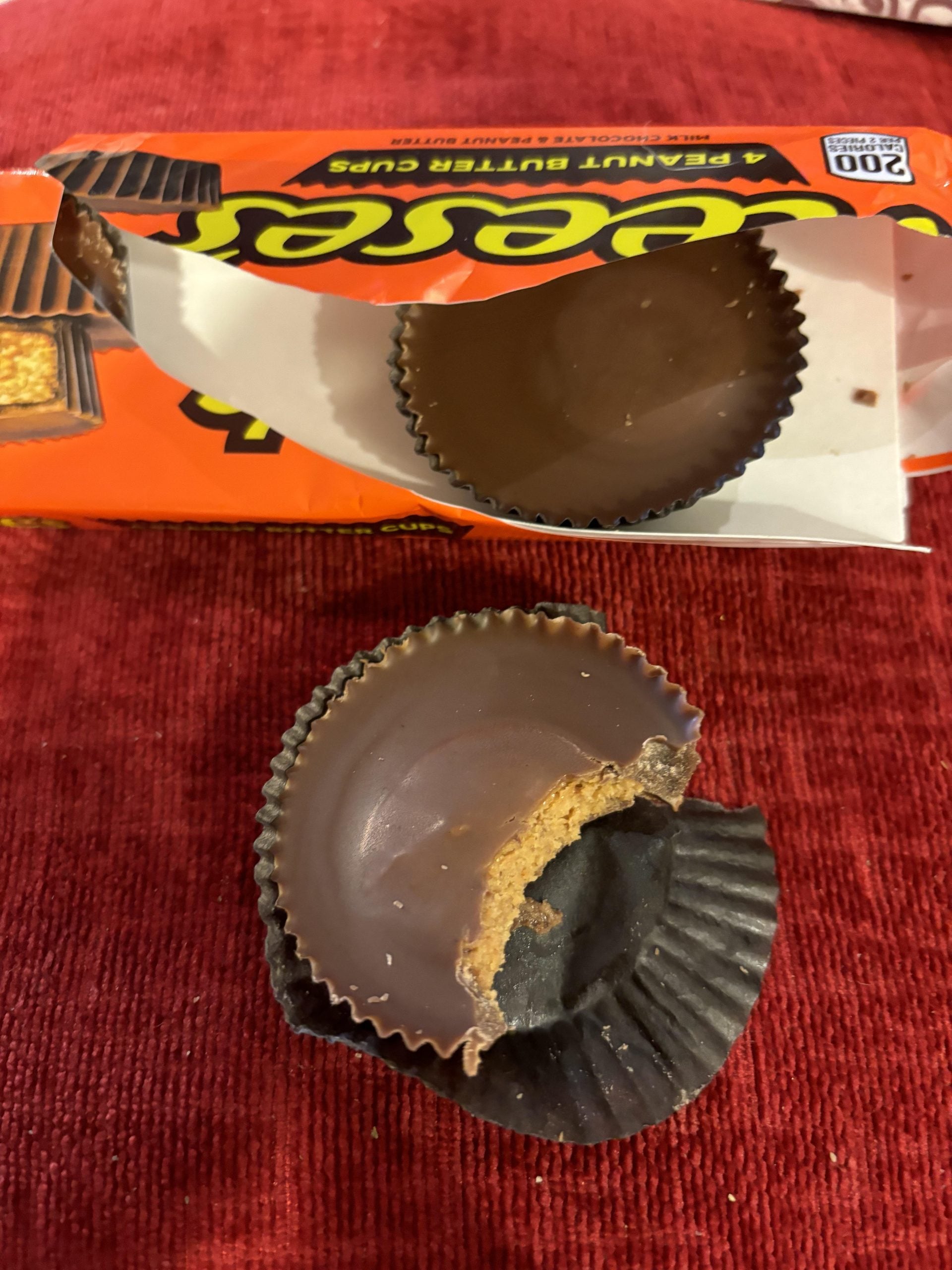 A Reese’s Peanut Butter Cup package with two cups, one bitten, resting on a red surface.