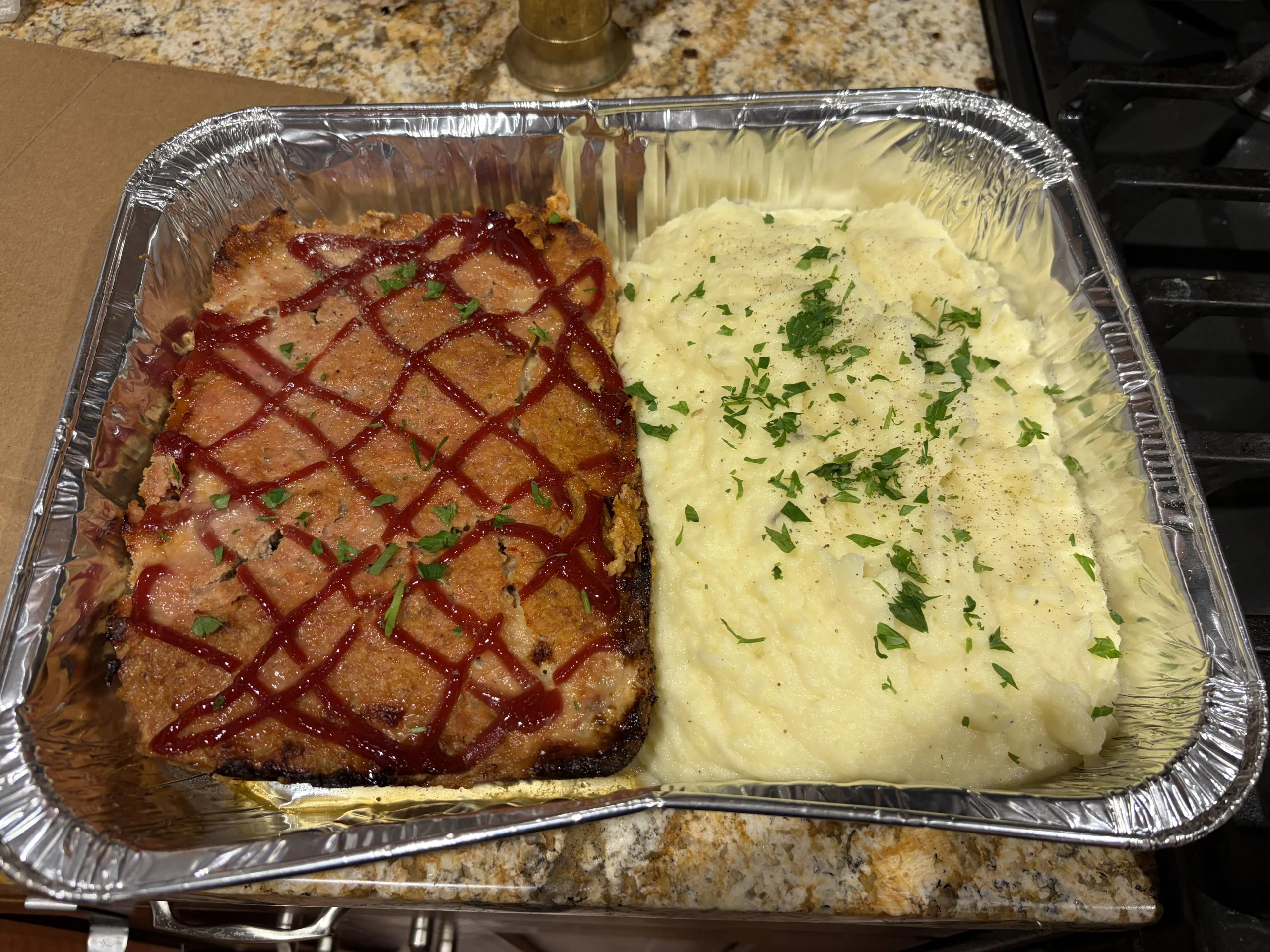 A foil tray with meatloaf topped with ketchup next to a serving of mashed potatoes garnished with chopped parsley, set on a granite kitchen countertop.