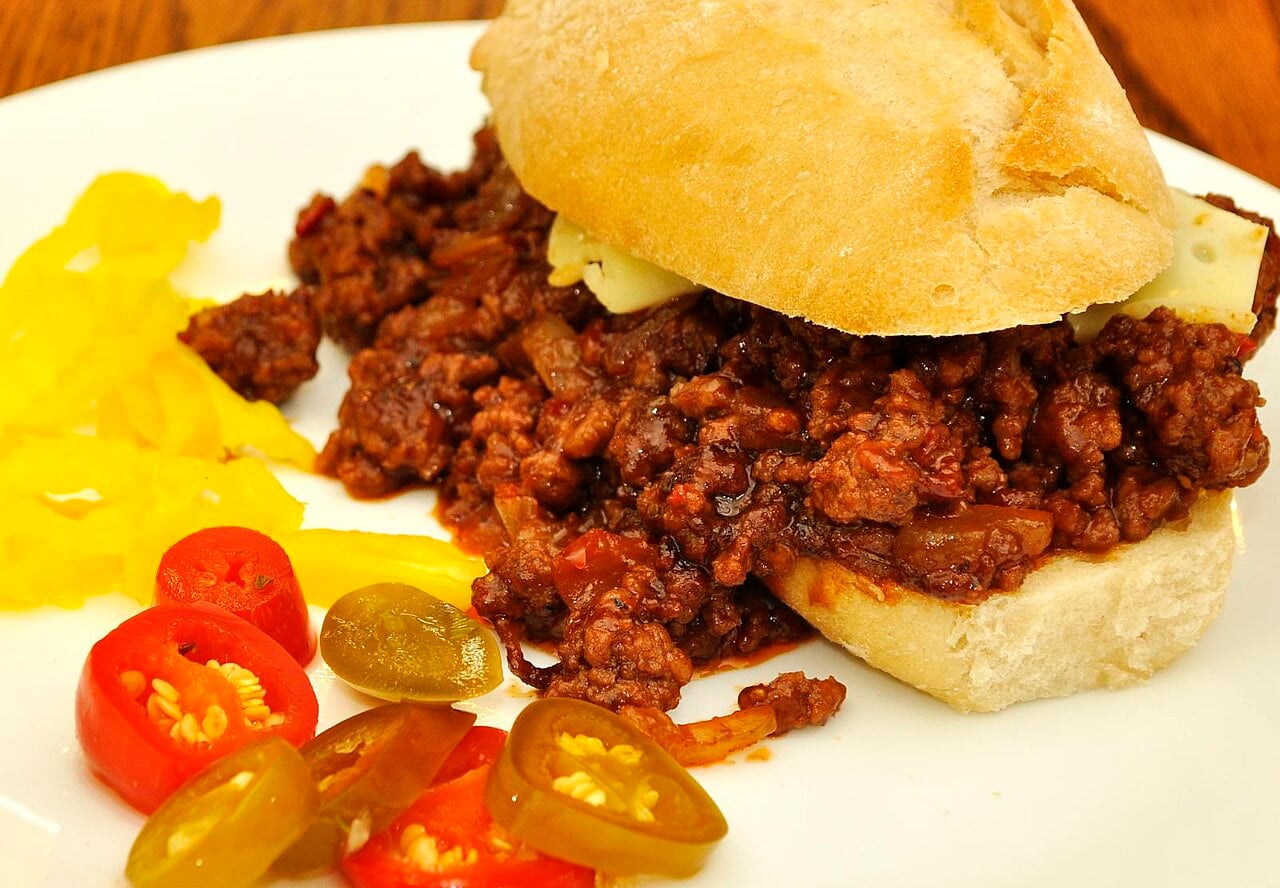 A sloppy joe sandwich on a white bun with a side of sliced yellow peppers and red chili peppers.