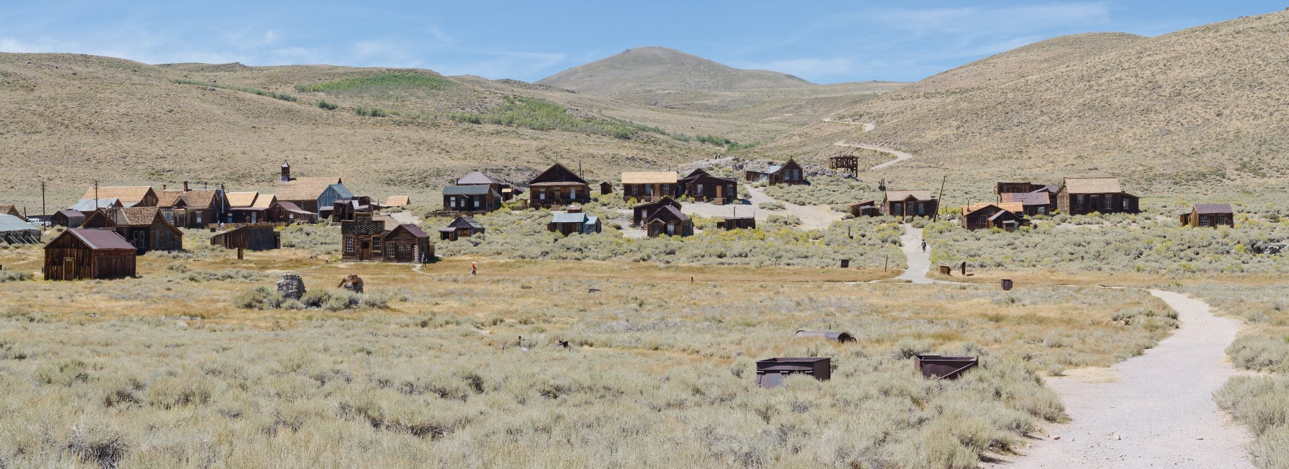 A panoramic view of the Bodie ghost town, featuring numerous weathered wooden buildings scattered across a dry, hilly landscape under a clear blue sky.
