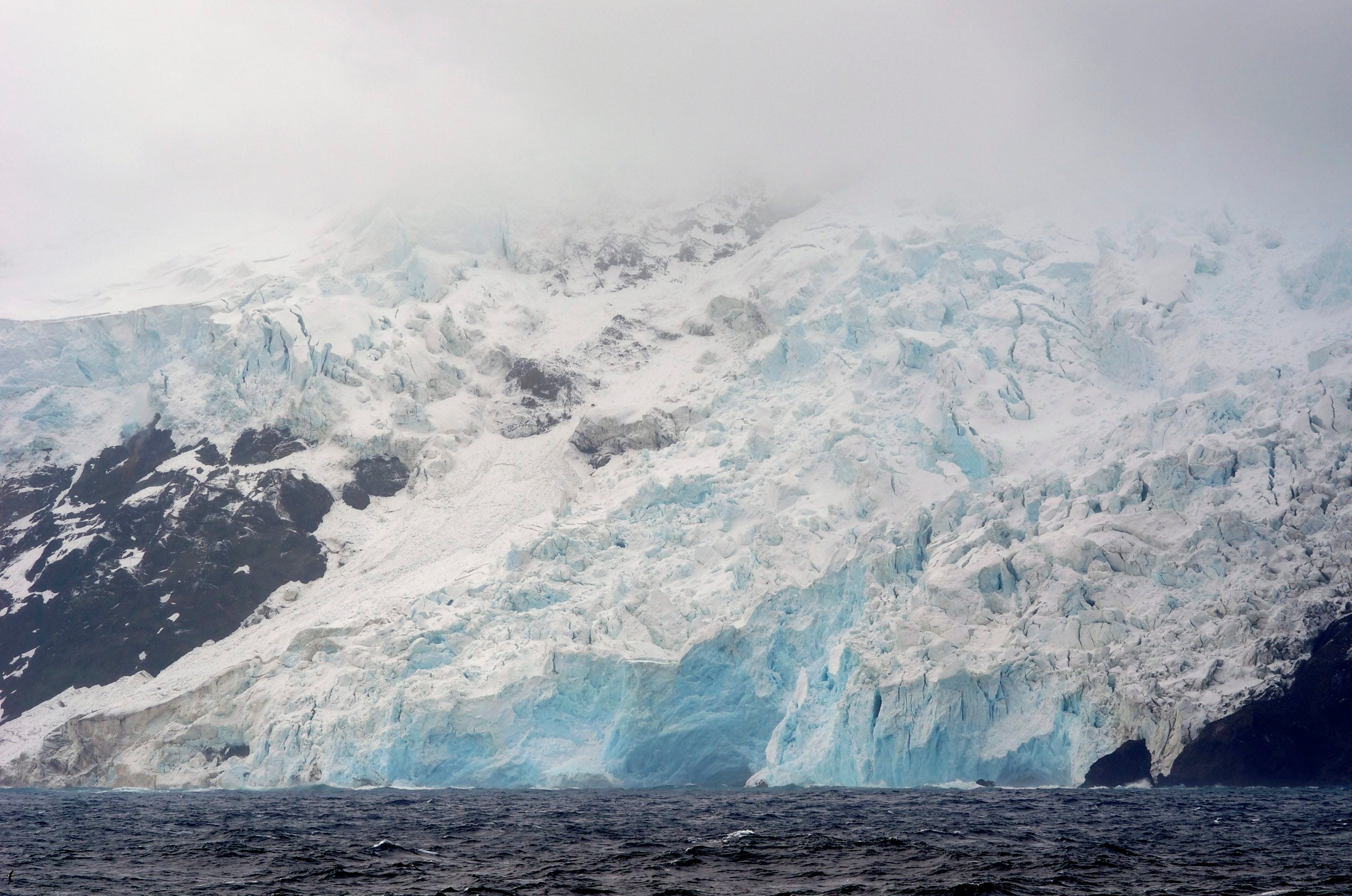 A massive glacier with deep blue ice shelves meets the dark, choppy ocean beneath a misty, cloud-covered mountain peak.