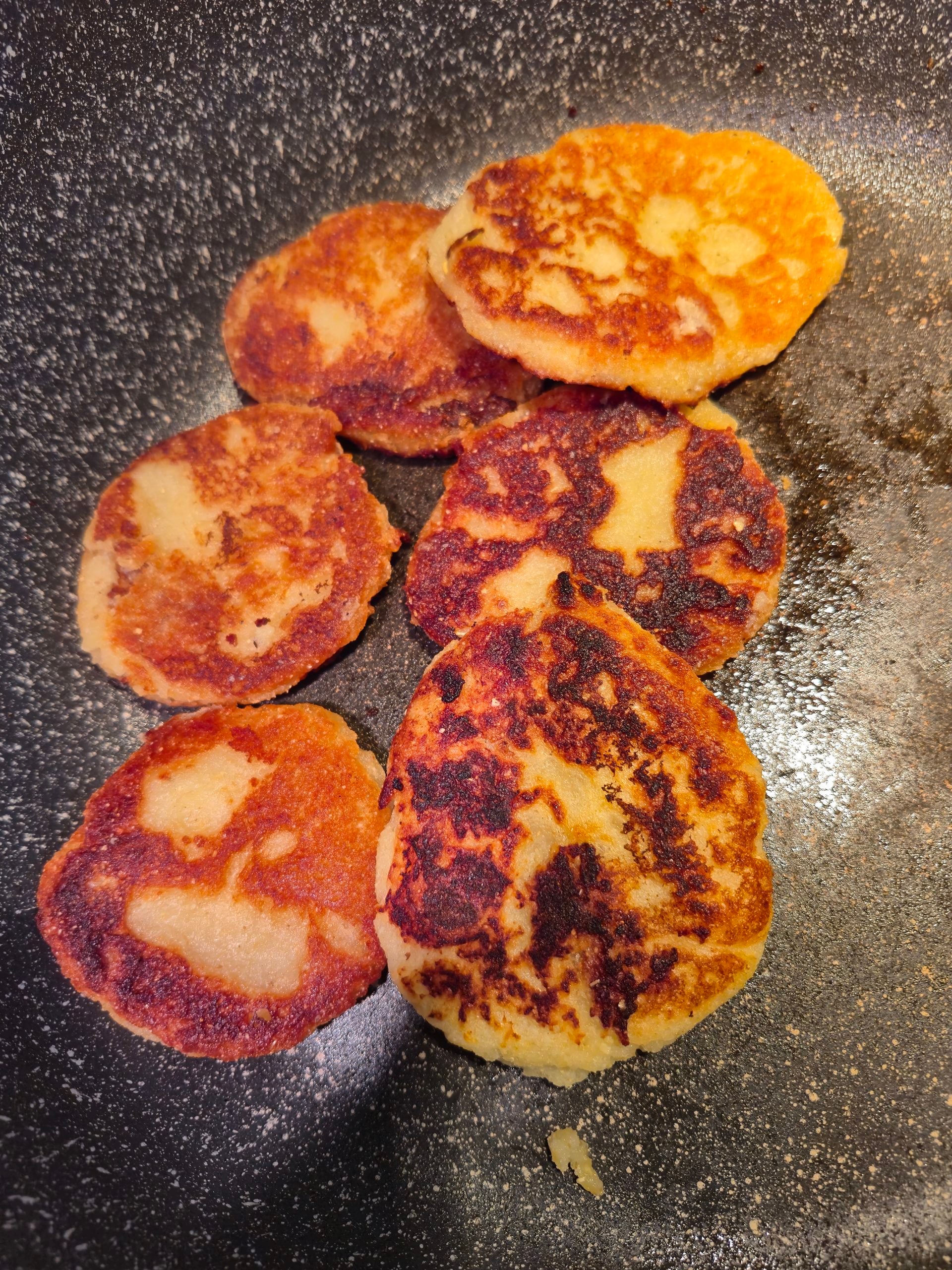 Six golden-brown potato pancakes cooking in a nonstick pan, showing crispy edges and varied browning on their surfaces.