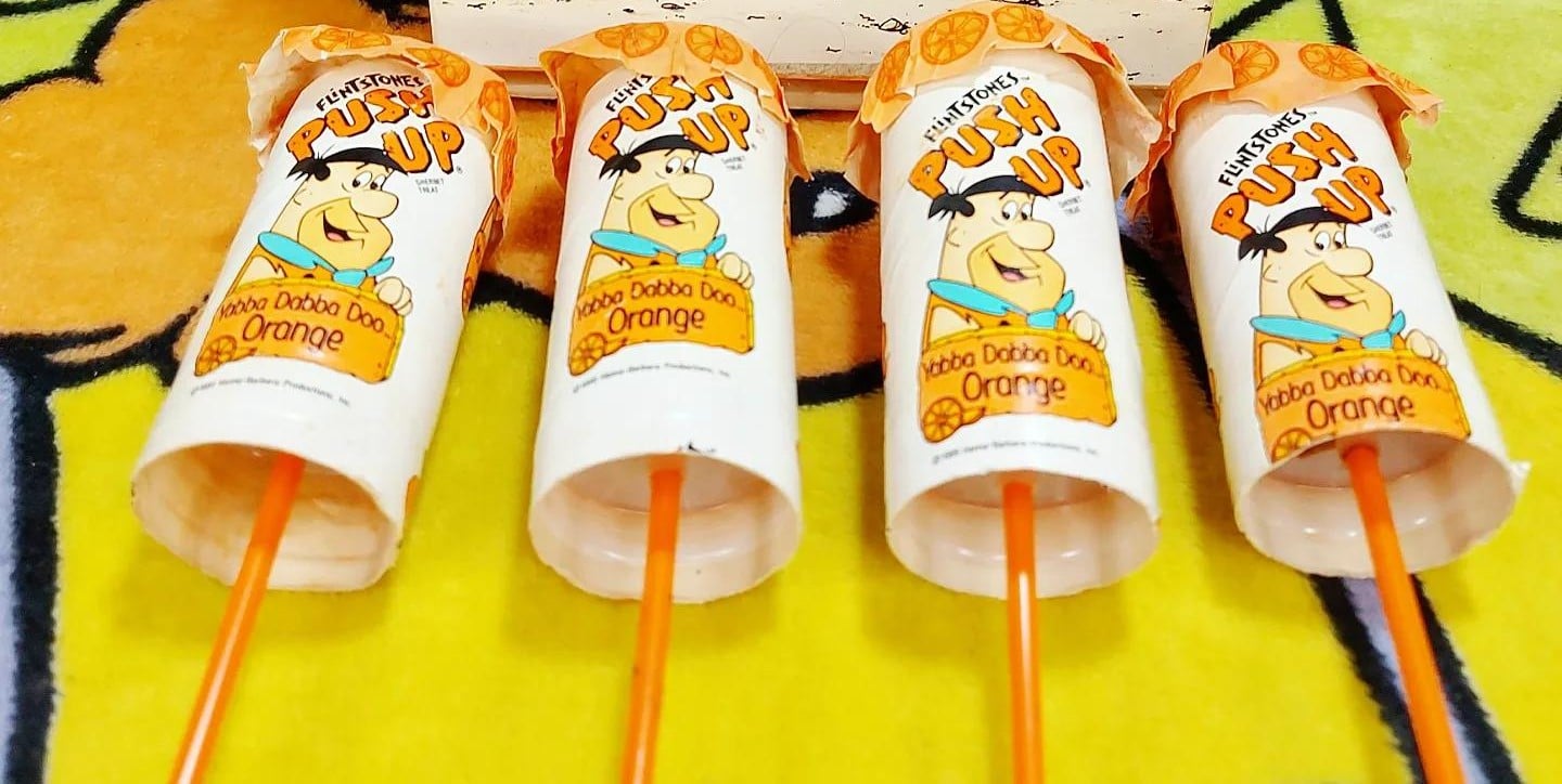 Four Flintstones orange Push-Up ice cream treats standing in a row on a yellow patterned surface.