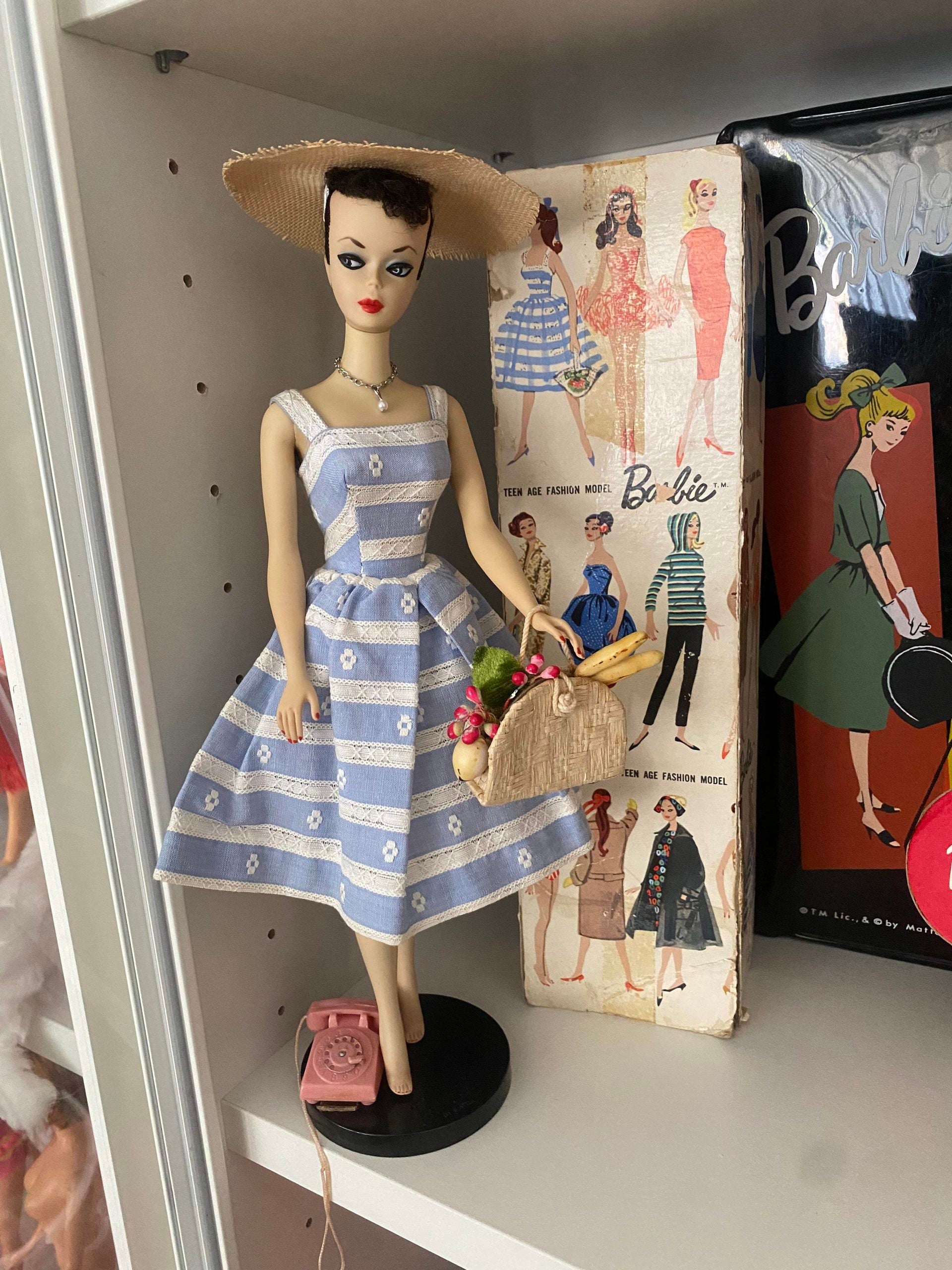 Vintage Barbie doll in a blue and white striped dress and straw hat, holding a basket with flowers. She stands on a shelf next to retro Barbie boxes and a pink toy phone.
