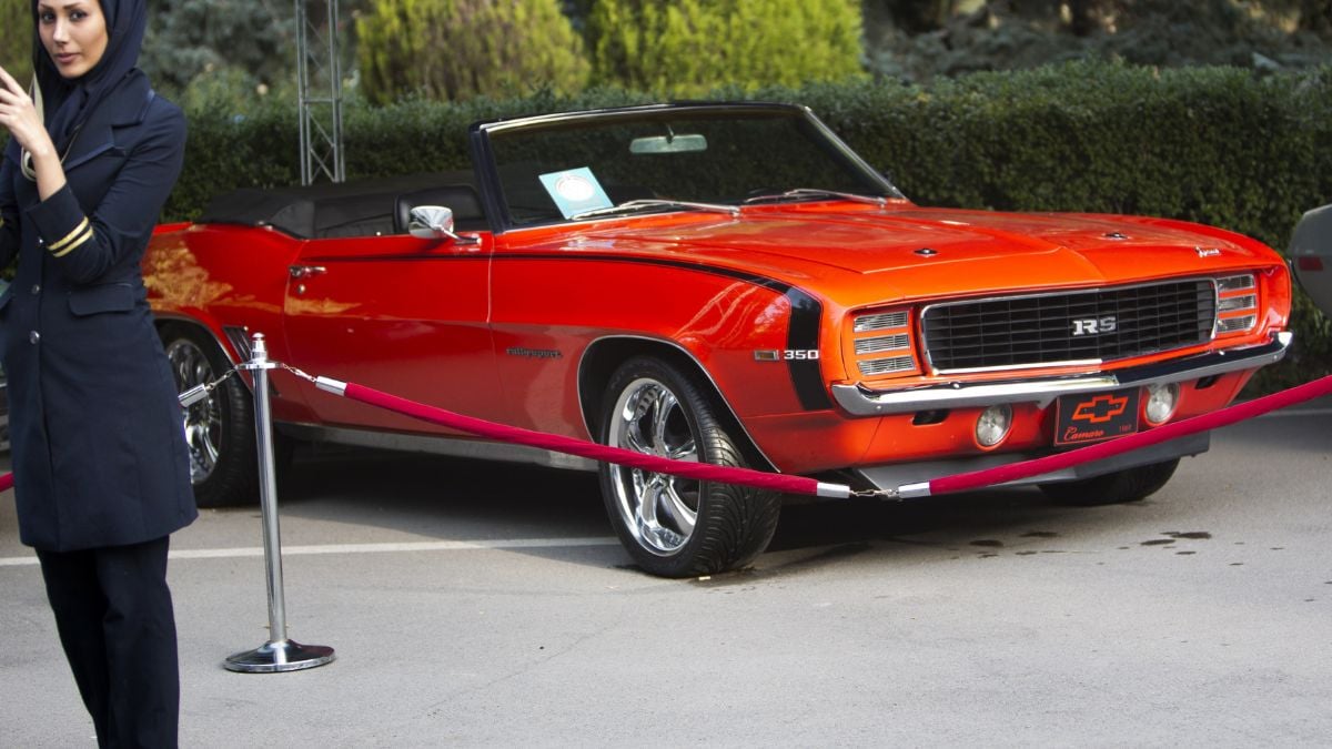 A bright orange classic Chevrolet Camaro RS convertible is displayed outdoors behind a red rope barrier. A person in dark clothing stands partially in view on the left side of the image.