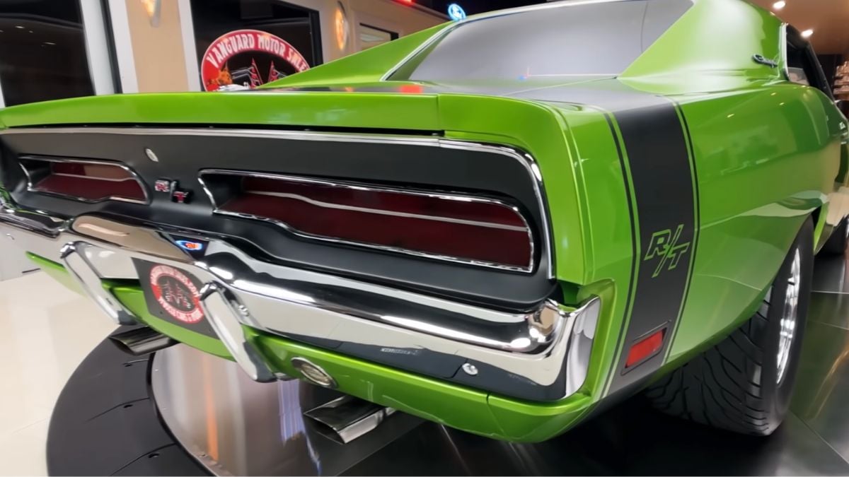 A close-up of the rear end of a green classic Dodge Charger R/T, showing its taillights, black racing stripes, chrome bumper, and wide tires, displayed indoors under bright lights.