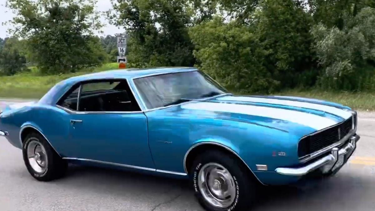 A classic blue muscle car with white racing stripes is parked on a road near trees and greenery, with a route sign visible in the background.