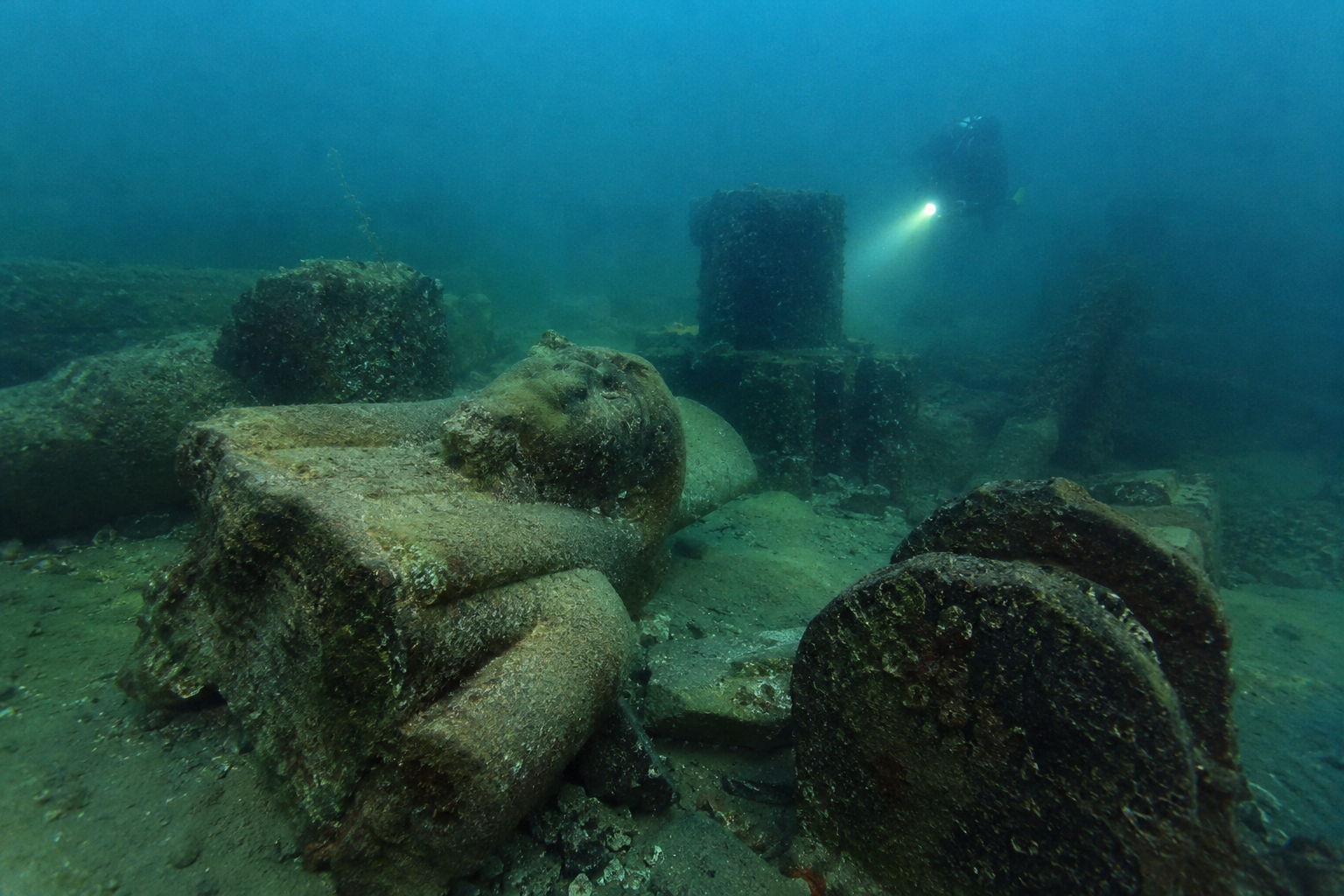 A scuba diver explores ancient stone ruins submerged on the ocean floor, including a partial statue of a human figure.