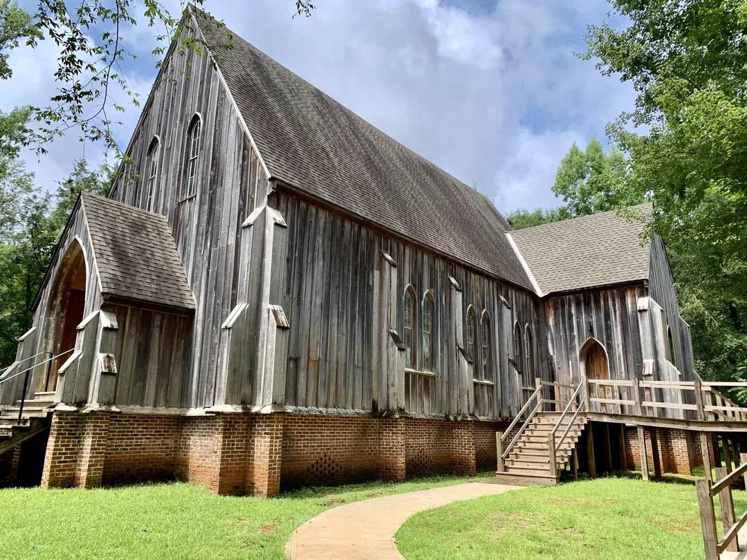 A wooden church with a steep roof and a brick foundation, surrounded by trees with a concrete path leading to wooden stairs.