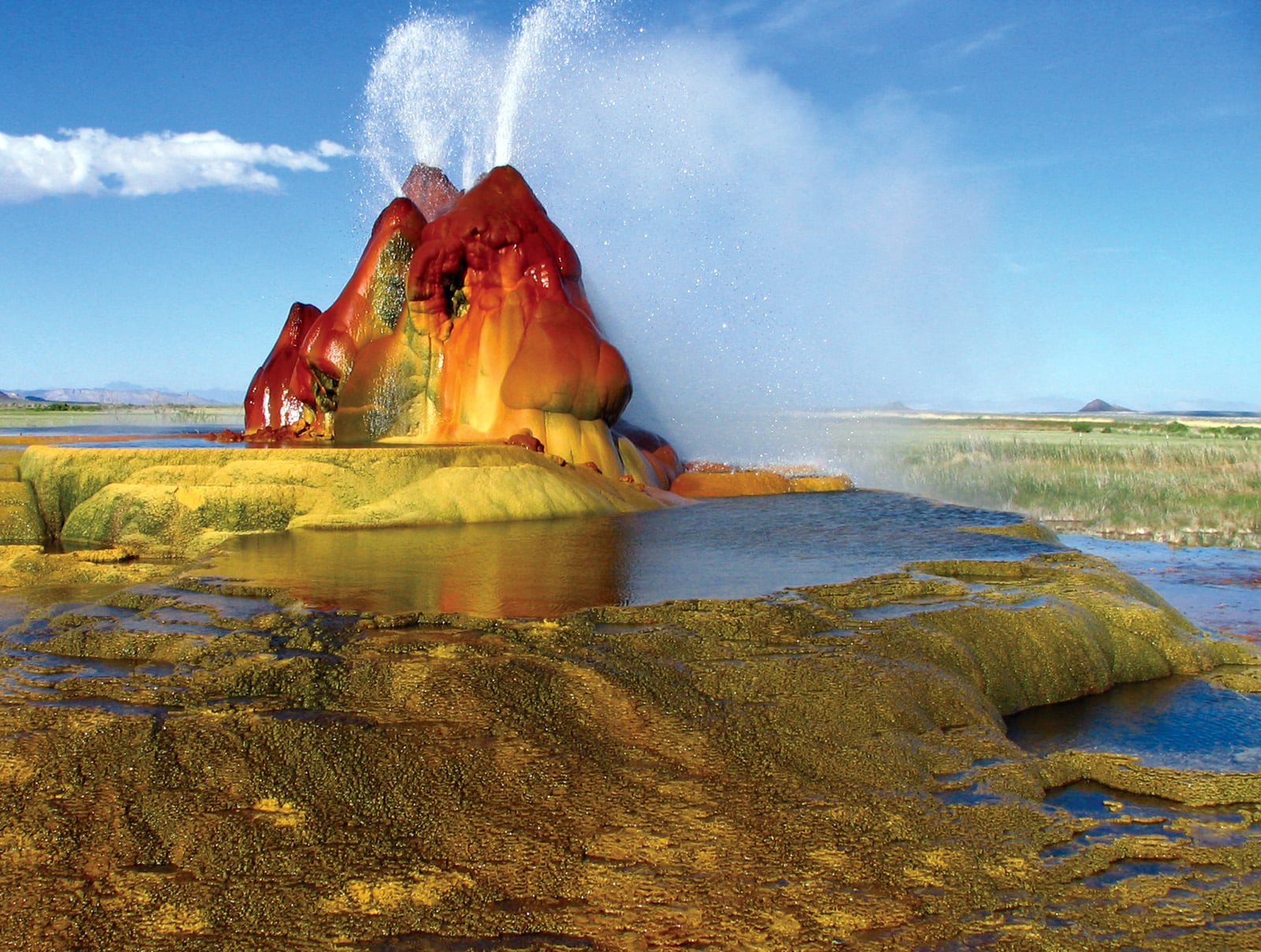 A colorful geothermal geyser erupts from a textured, mineral-rich mound in a grassy field.