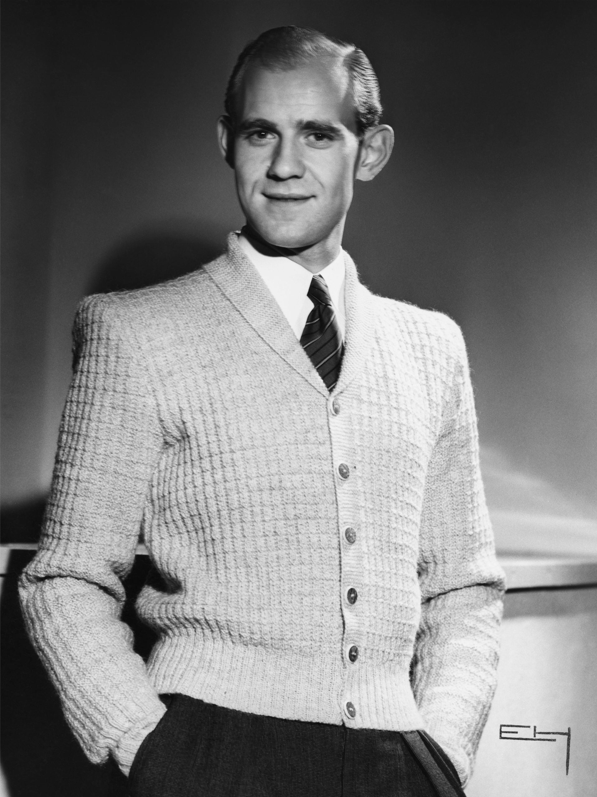 A man wearing a light-colored, buttoned cardigan over a shirt and tie stands with hands in pockets, smiling slightly. The image is black and white and appears to be from a mid-20th century photo shoot.