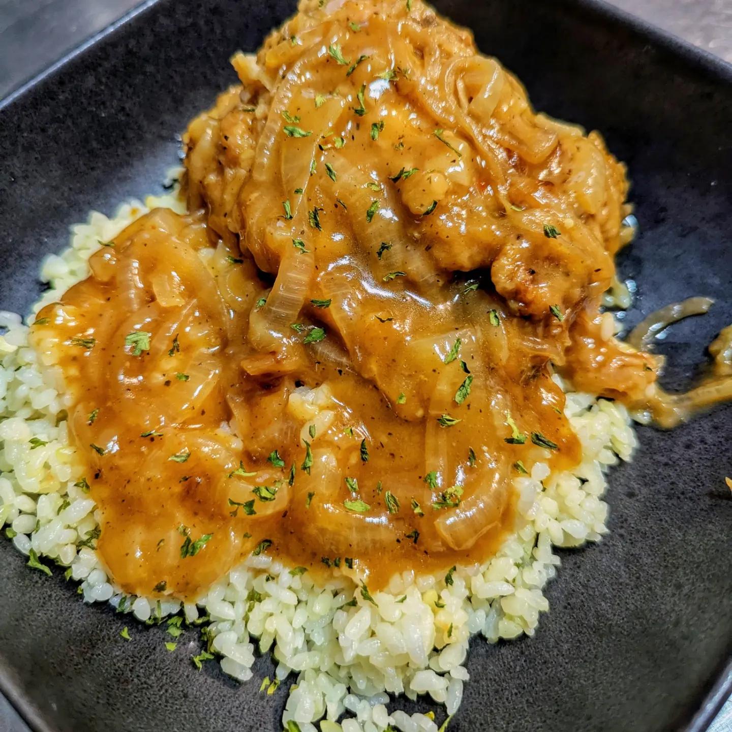 Chicken with brown onion gravy served over a bed of rice, garnished with herbs, on a black square plate.