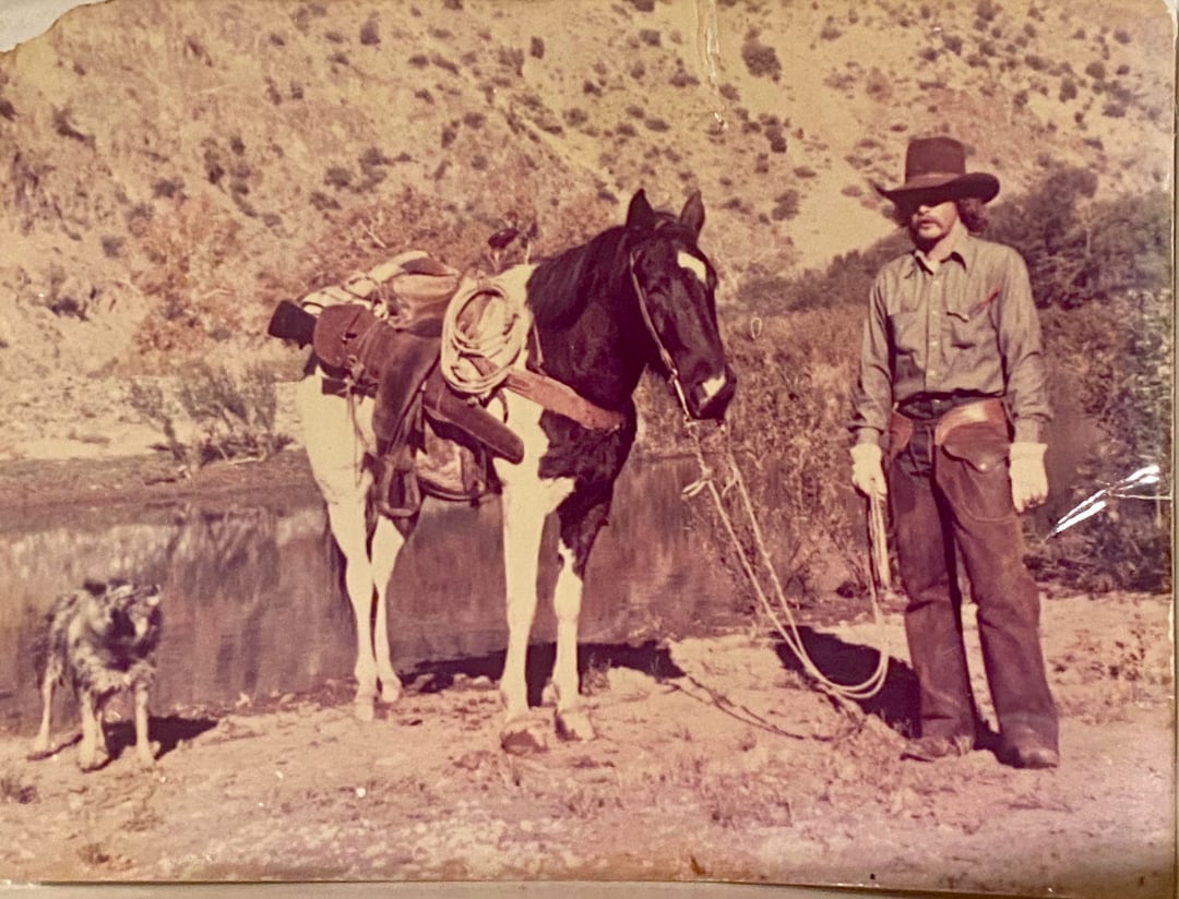 A cowboy in a hat and chaps stands next to his saddled horse by a river, with a dog standing nearby.