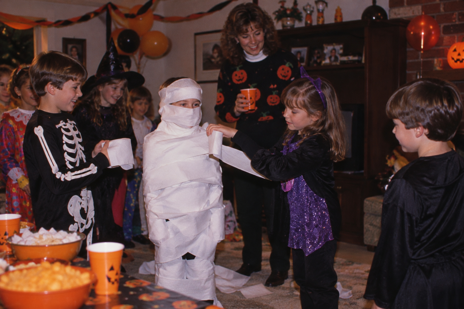 Children in Halloween costumes wrapping a friend in toilet paper to look like a mummy at a party.
