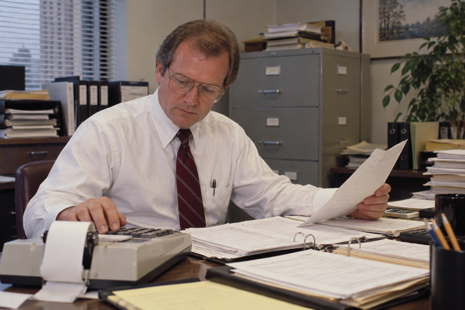 A man in a white dress shirt and tie sitting at an office desk while reviewing documents and using a printing calculator.
