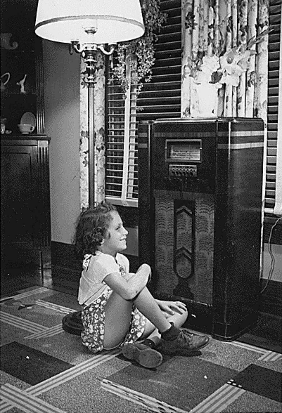 A young girl sits on the floor smiling at a large vintage radio console in a living room.