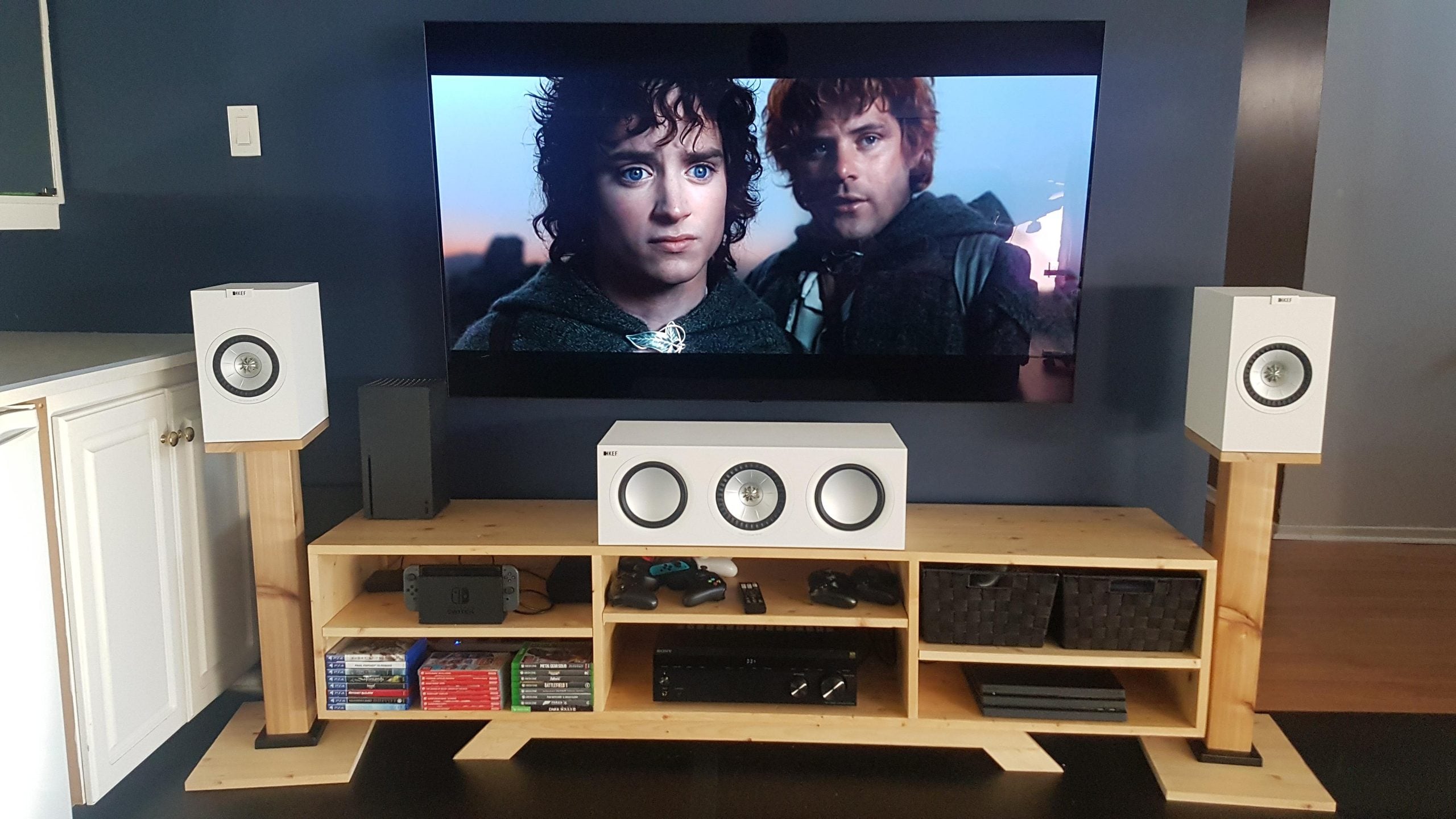 A television mounted on a wall above a wooden media console, displaying a scene from The Lord of the Rings, with two white bookshelf speakers on wooden stands and a white center channel speaker on the console.