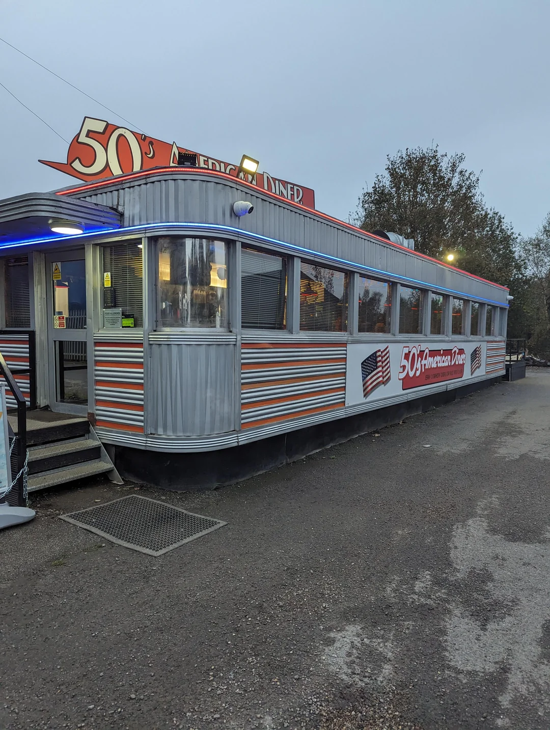 A retro-style 50s American Diner featuring a corrugated metal exterior, blue neon lights, and a sign displaying the name with an American flag.