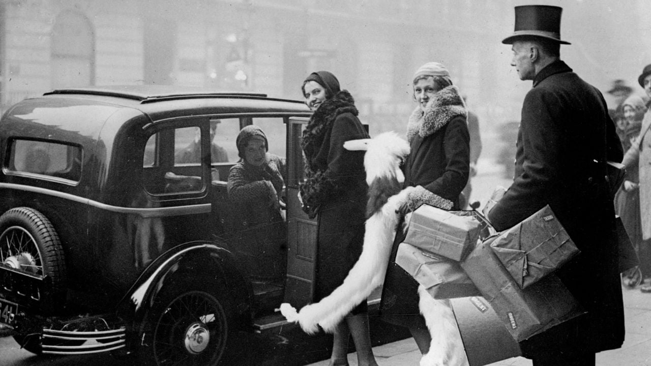 Three women in 1920s attire stand by a vintage car, one seated inside. A man in a top hat holds packages nearby, all dressed warmly on a city street.