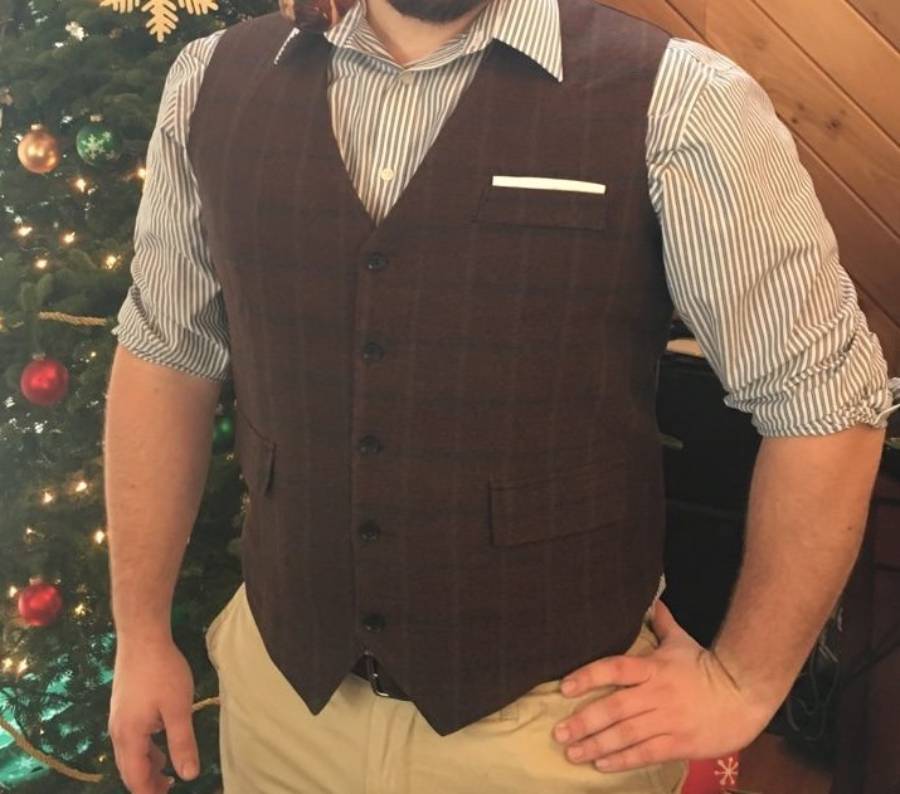 A person wearing a striped shirt, brown vest, and khaki pants stands with one hand on their hip in front of a decorated Christmas tree.