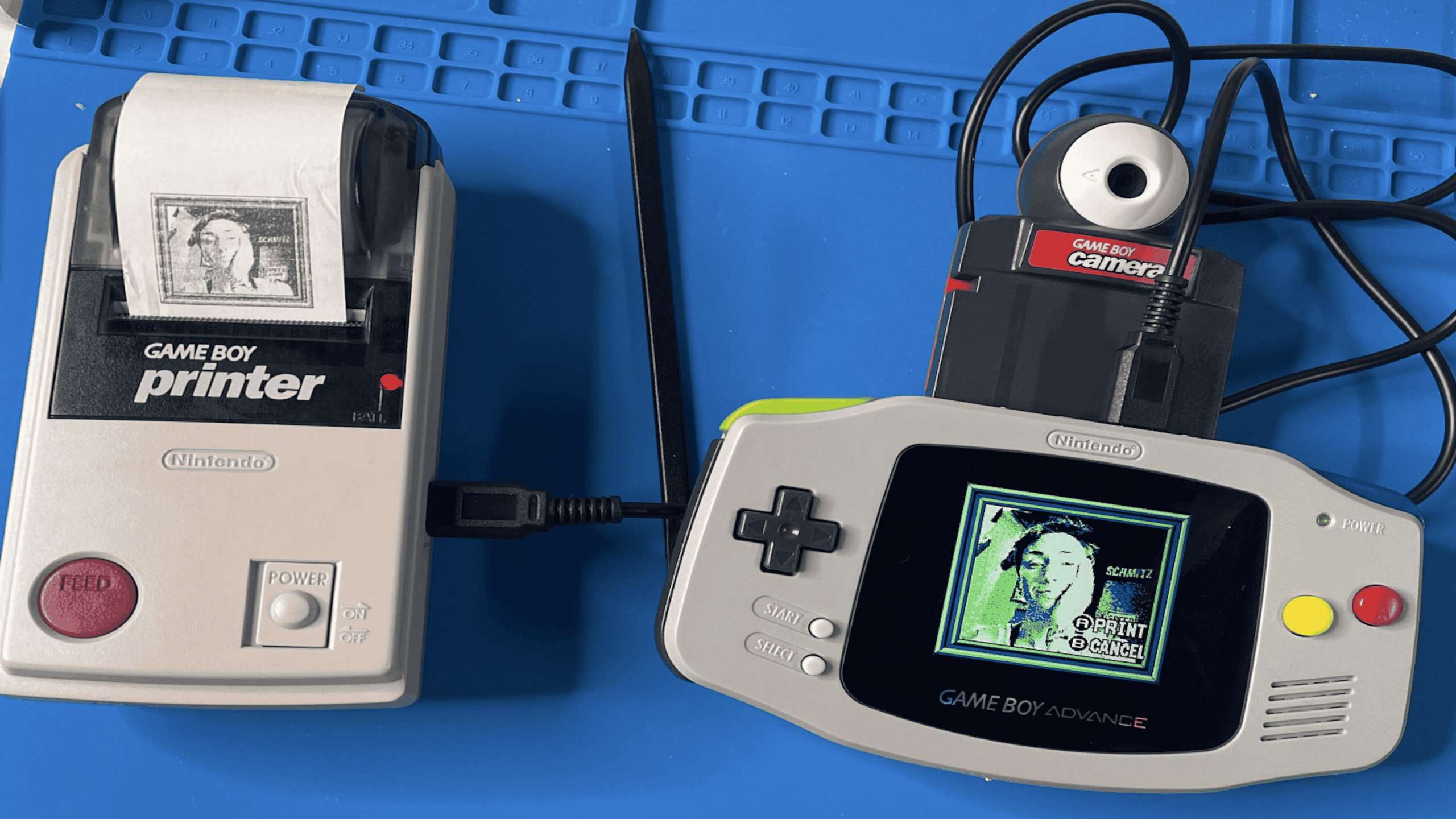 A Game Boy Advance with a Game Boy Camera attached, connected by a link cable to a Game Boy Printer that is printing a captured image.