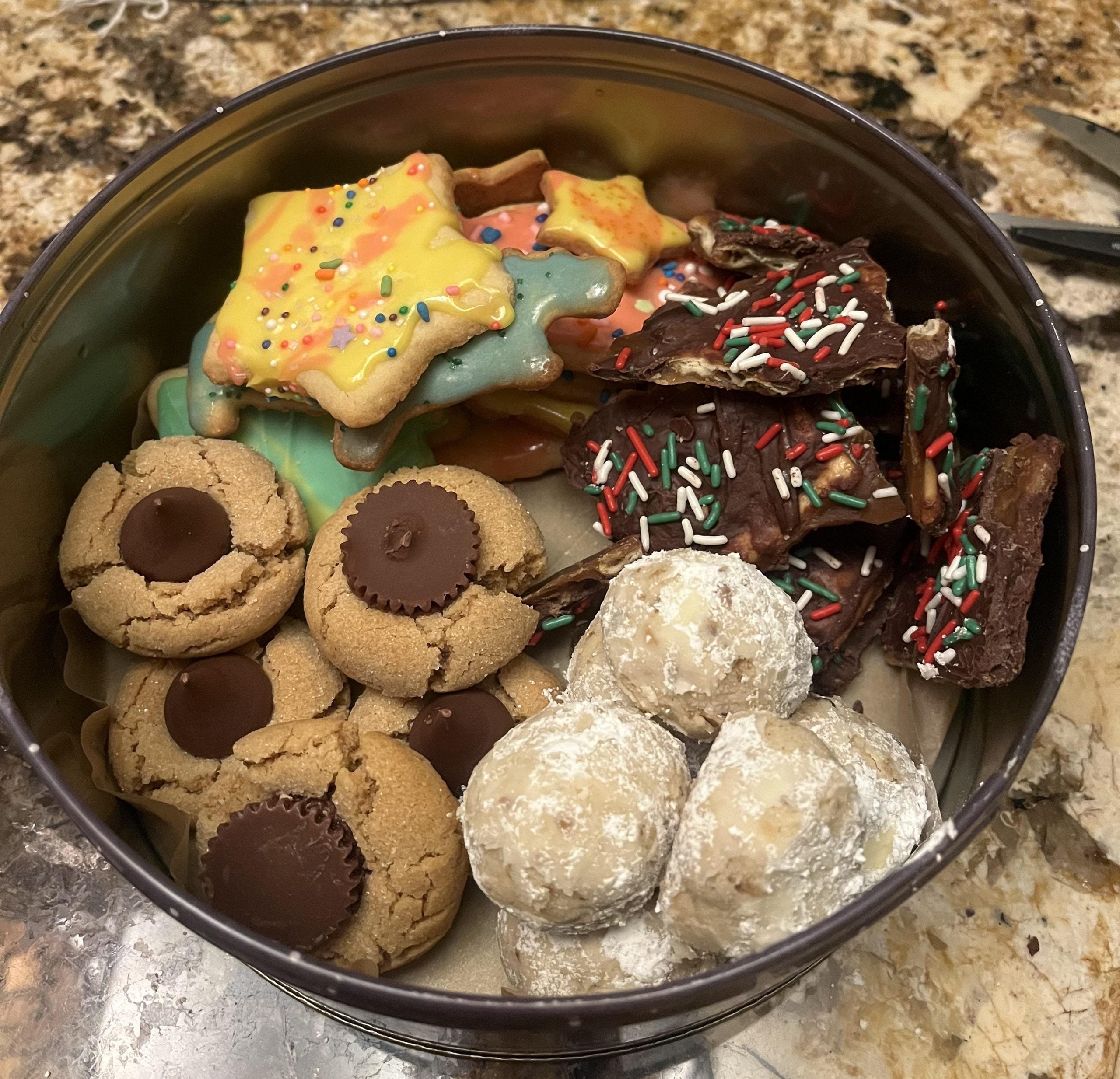 A tin filled with assorted holiday treats, including star-shaped sugar cookies, chocolate bark with sprinkles, peanut butter cup cookies, and powdered sugar-coated cookies.