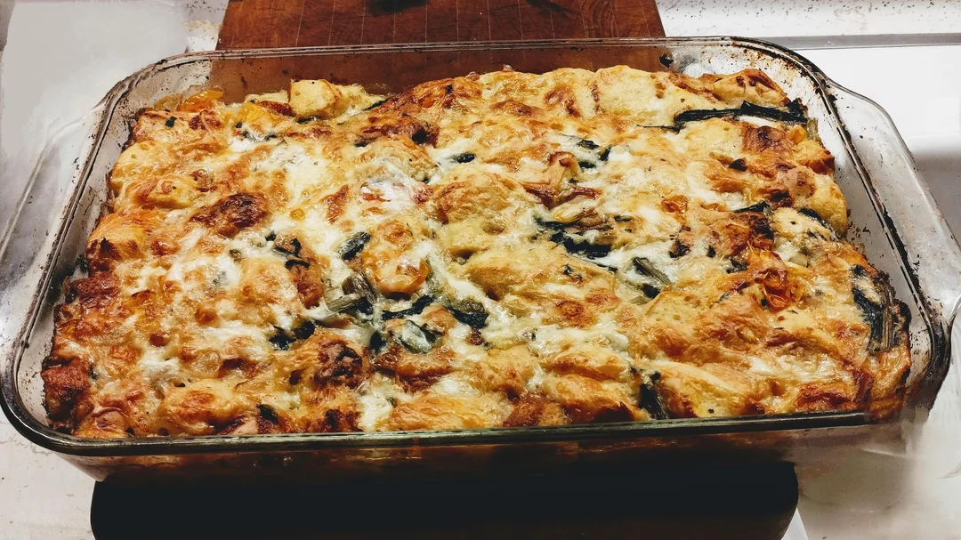 A glass baking dish filled with a golden-brown, cheesy casserole featuring visible pieces of bread, greens, and melted cheese on top, resting on a wooden board.