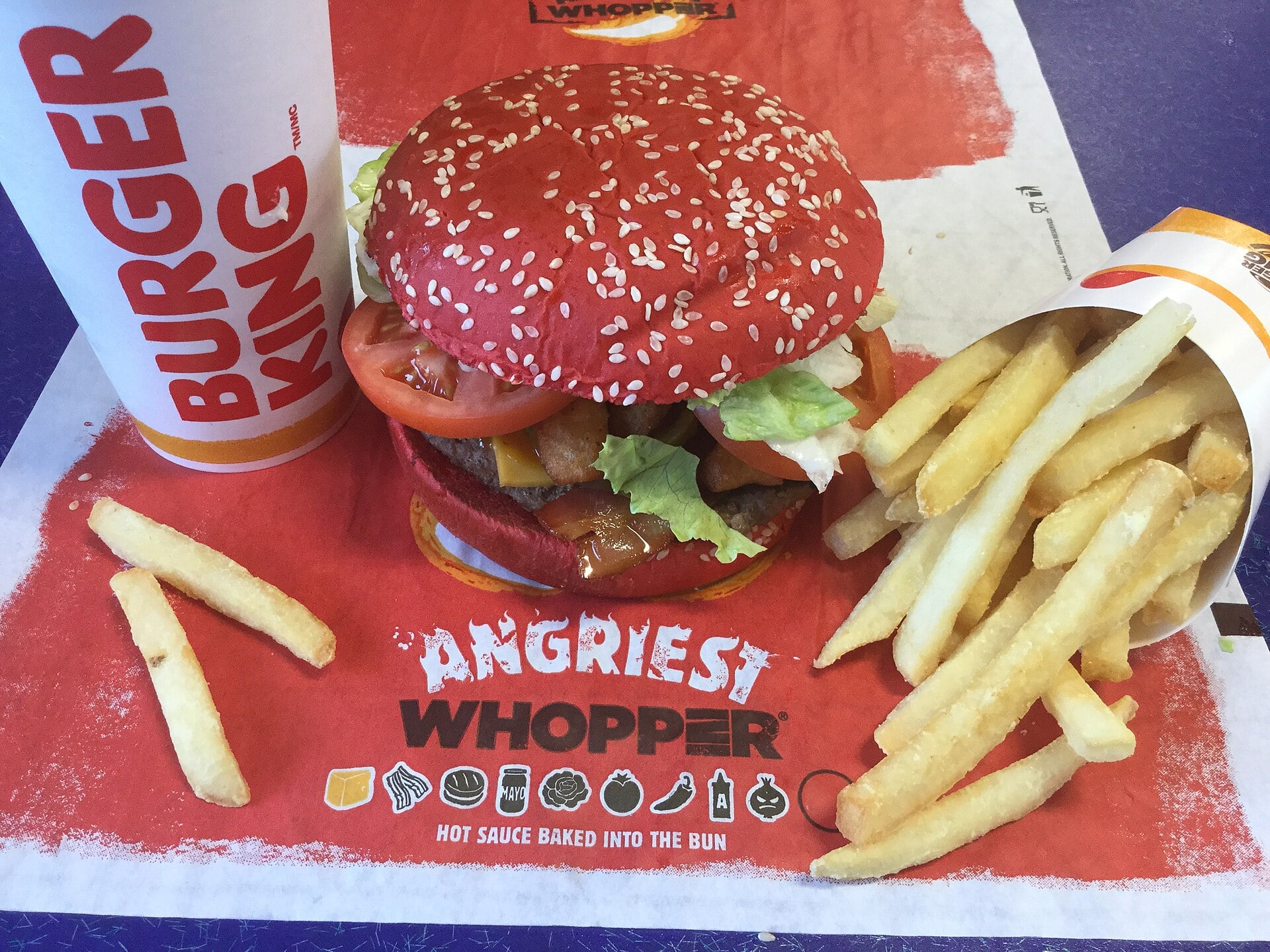 An Angriest Whopper with a red bun, a Burger King drink cup, and a carton of french fries on a branded tray liner.