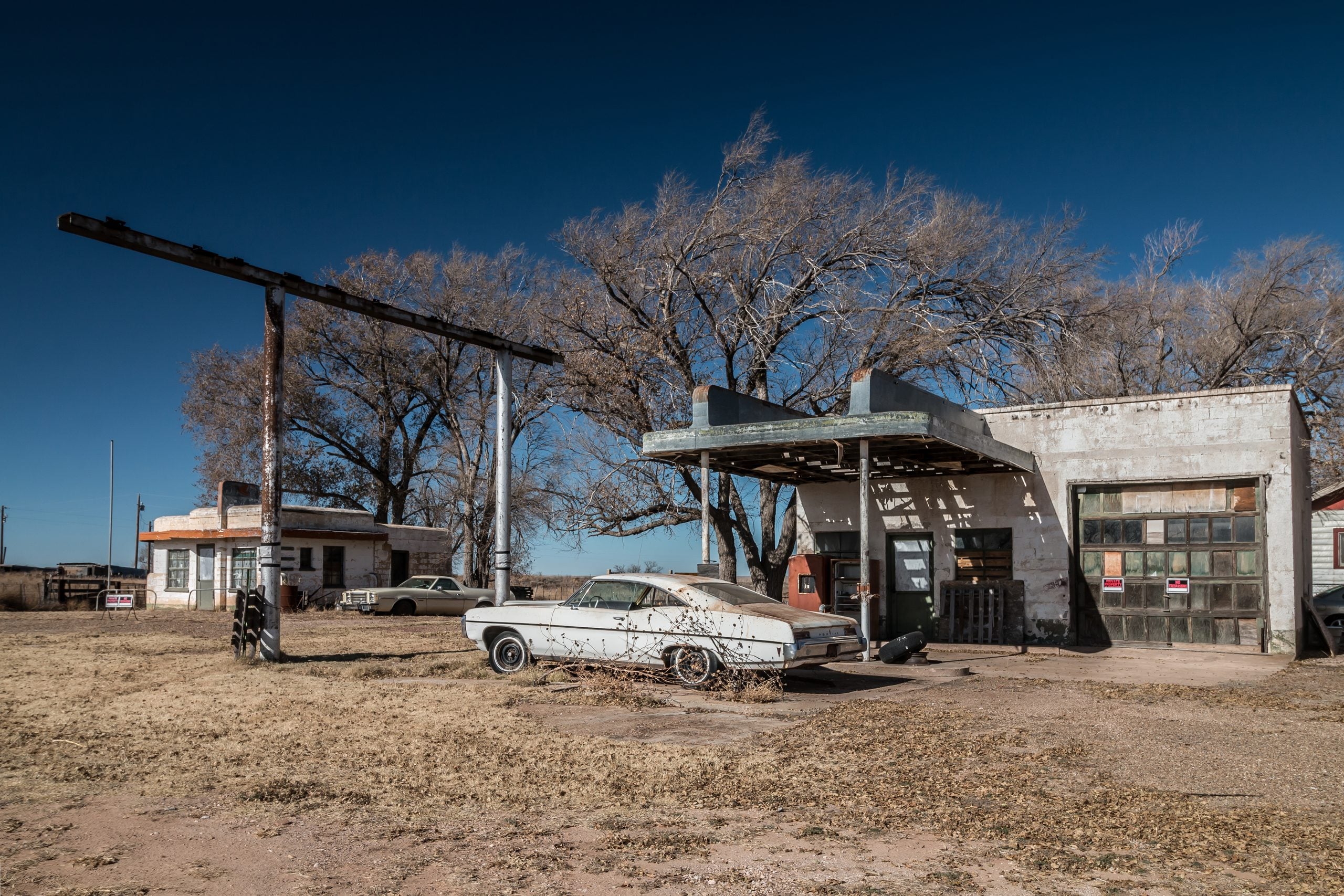 An abandoned gas station with a rusted vintage car parked outside, surrounded by dry grass and leafless trees under a clear blue sky. The buildings appear weathered and unused.
