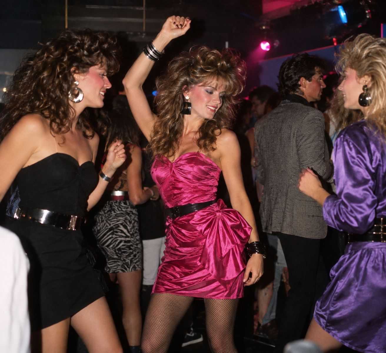Three women dancing at a nightclub, with one woman in the center wearing a bright pink strapless dress.