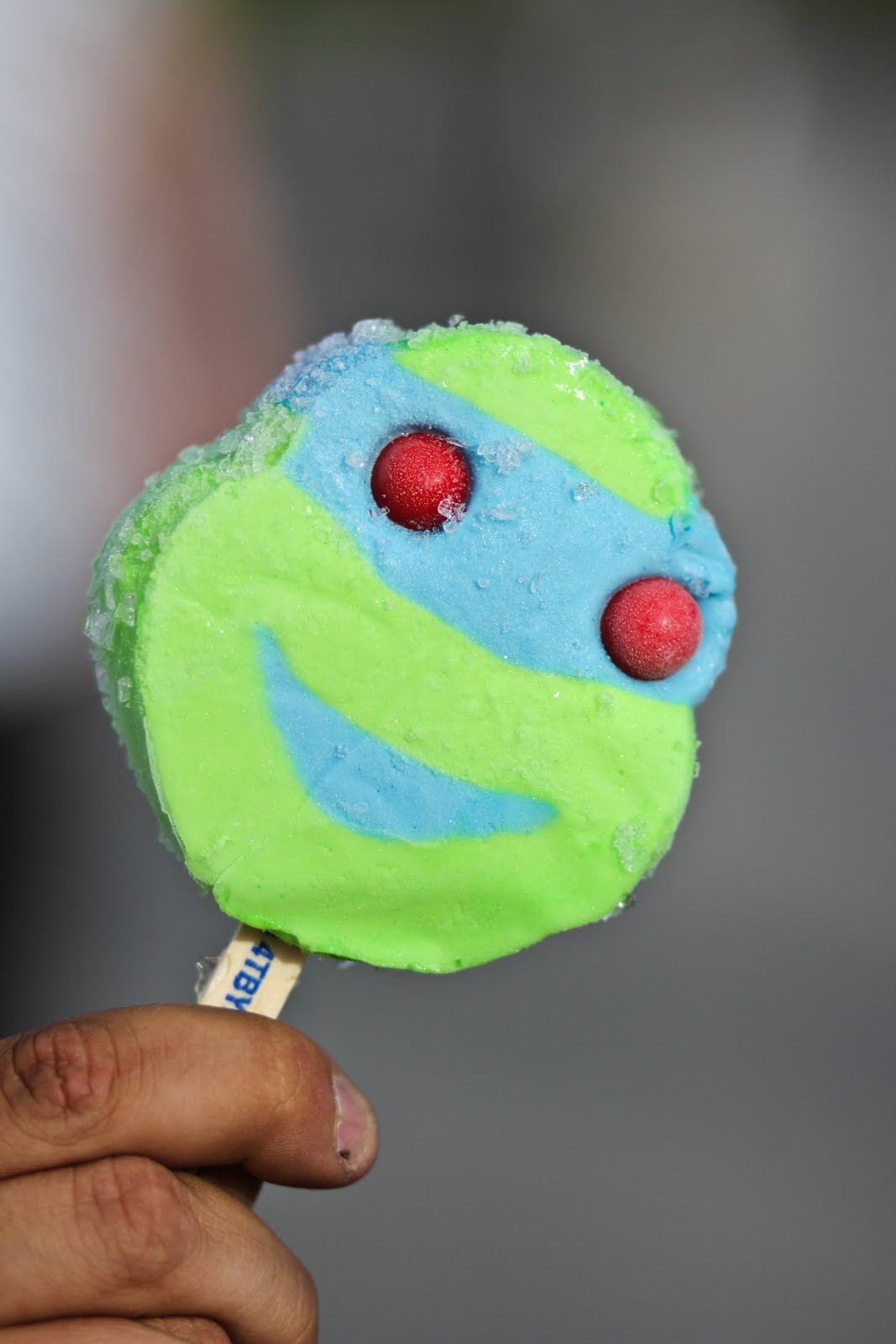 A hand holds a Teenage Mutant Ninja Turtles character popsicle with blue and green coloring and two red gumball eyes.