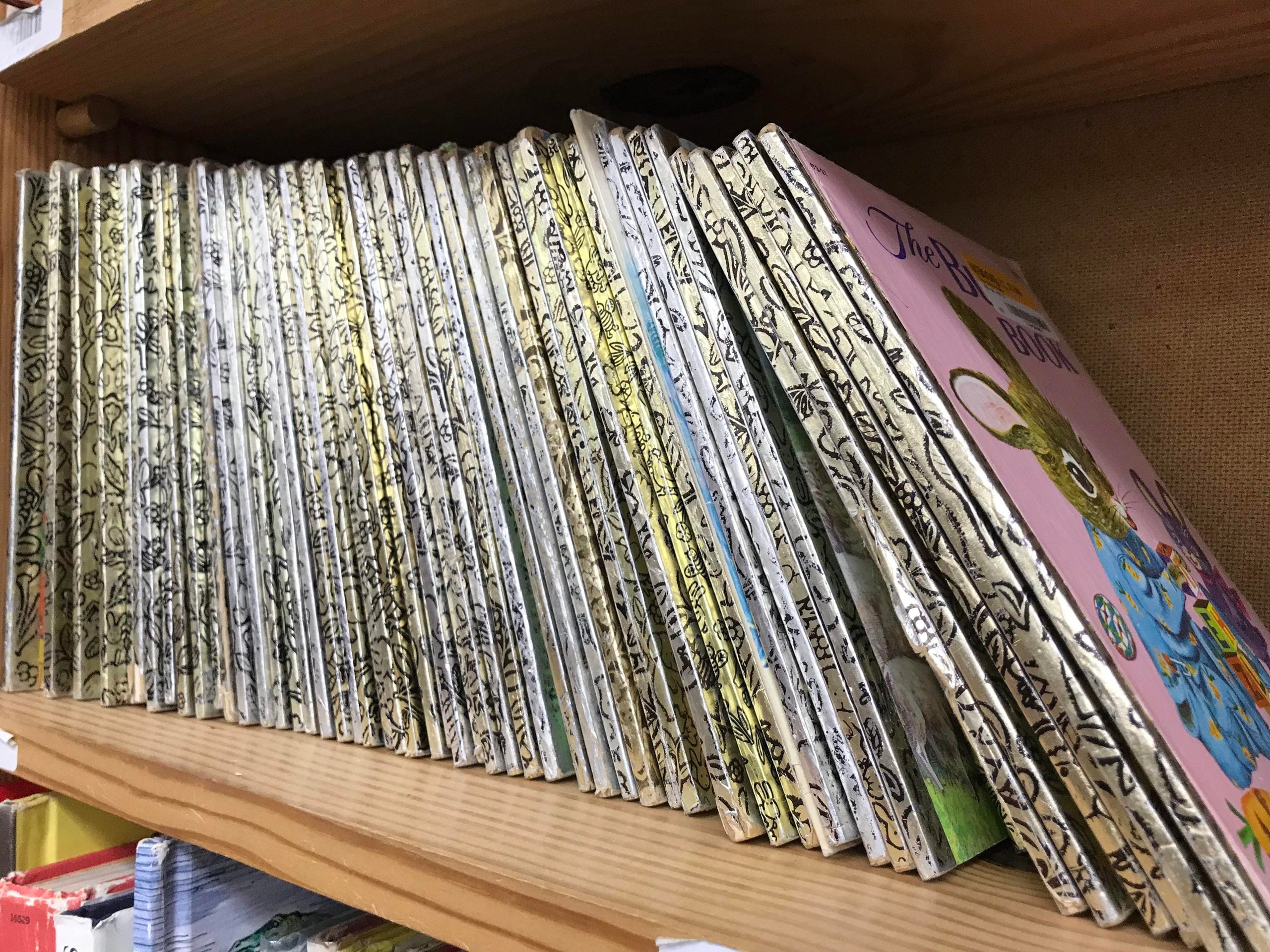 A long row of identical children’s books with gold-patterned spines lined up on a wooden shelf, with one book showing a pink cover featuring a rabbit illustration.