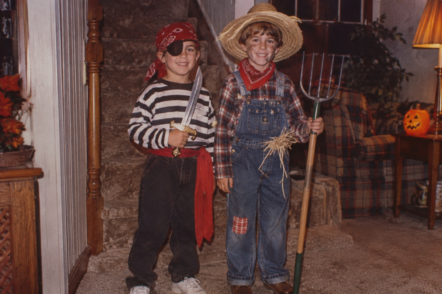 Two young boys in Halloween costumes, one dressed as a pirate with an eye patch and sword, and the other as a scarecrow wearing overalls and a straw hat while holding a pitchfork.